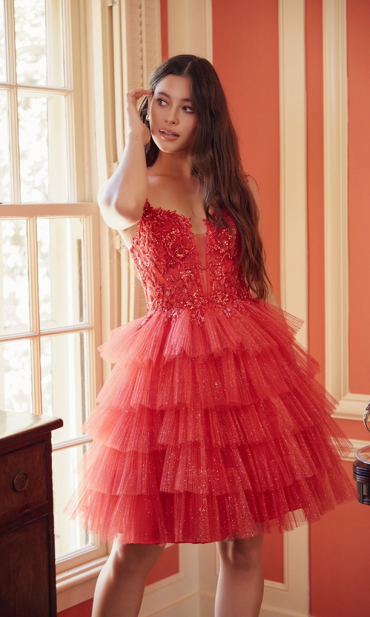 A woman stands indoors by a window, wearing the Glitter Babydoll Homecoming Dress: Nox Anabel E904, a tiered red corset dress with floral-embellished bodice, looking off to the side and touching her hair.